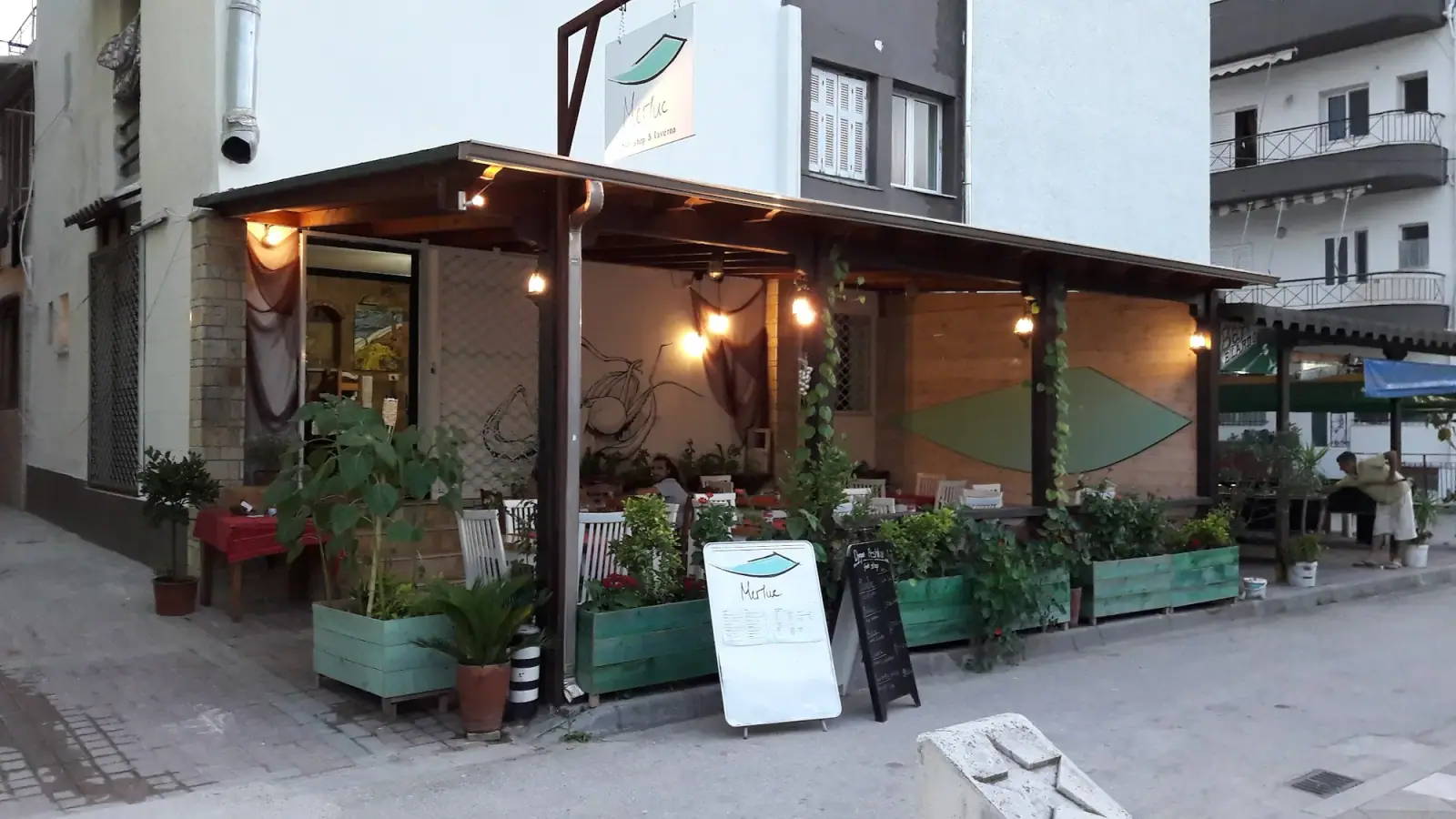 Covered terrace of Merluc in Spile at dusk, lamps lit above wooden planters and white chairs