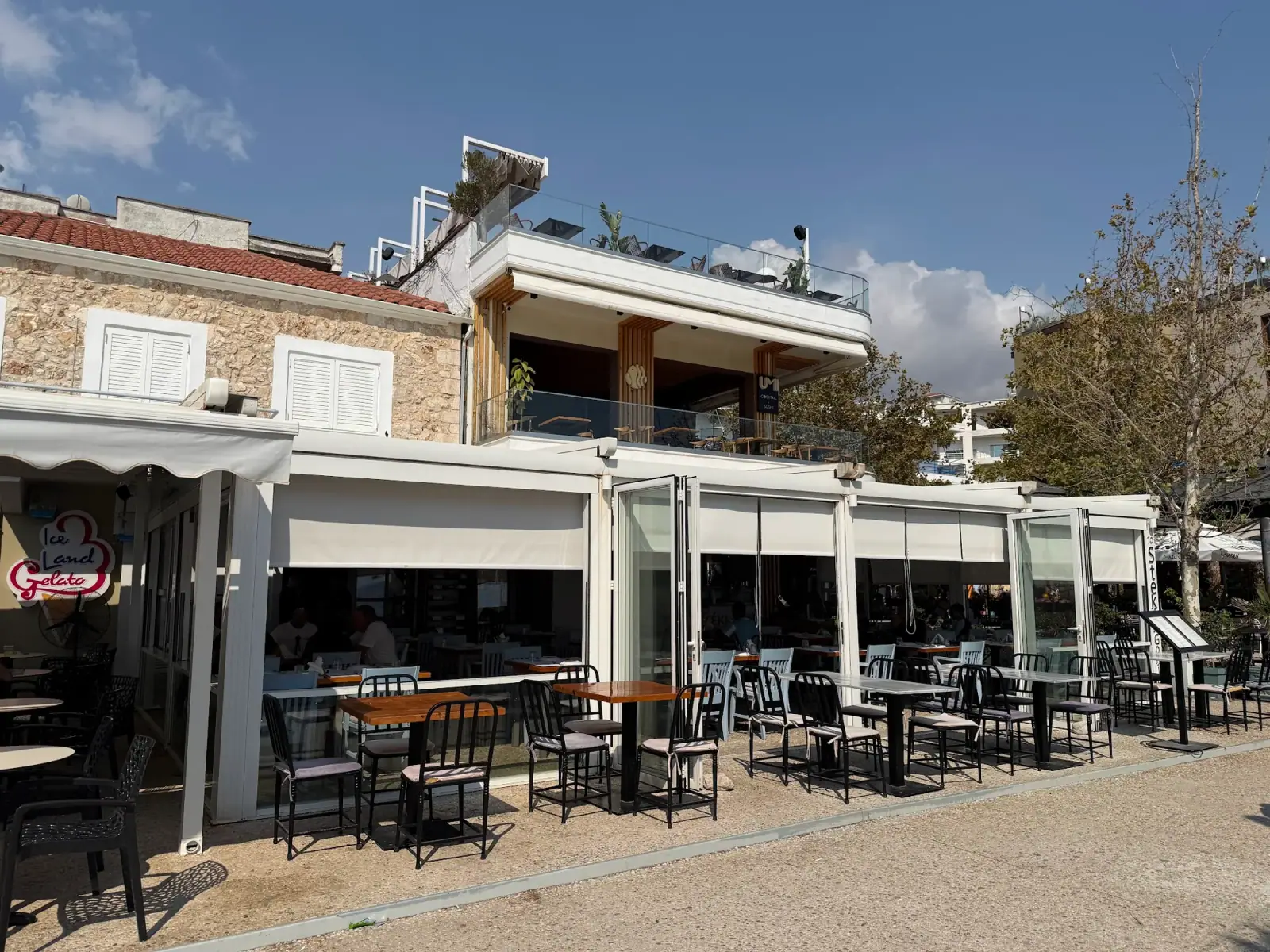 Outdoor tables in front of the folding glass walls of To Steki sti Gonia on the Spile promenade, Himarë