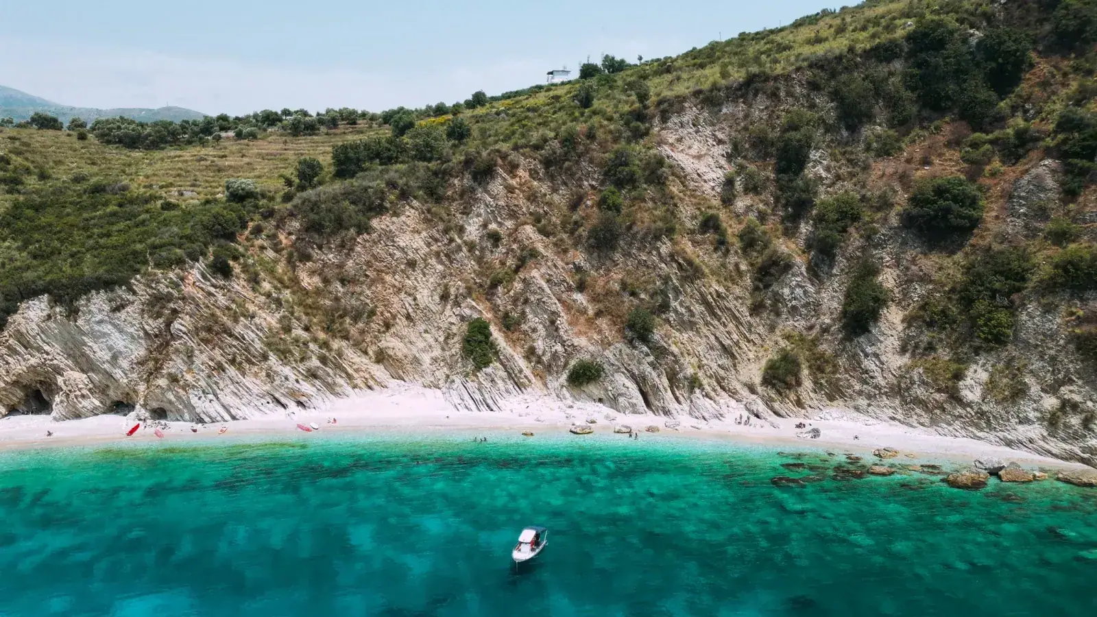 A white-pebble beach below limestone cliffs on the Himara coast, with a boat anchored in turquoise water