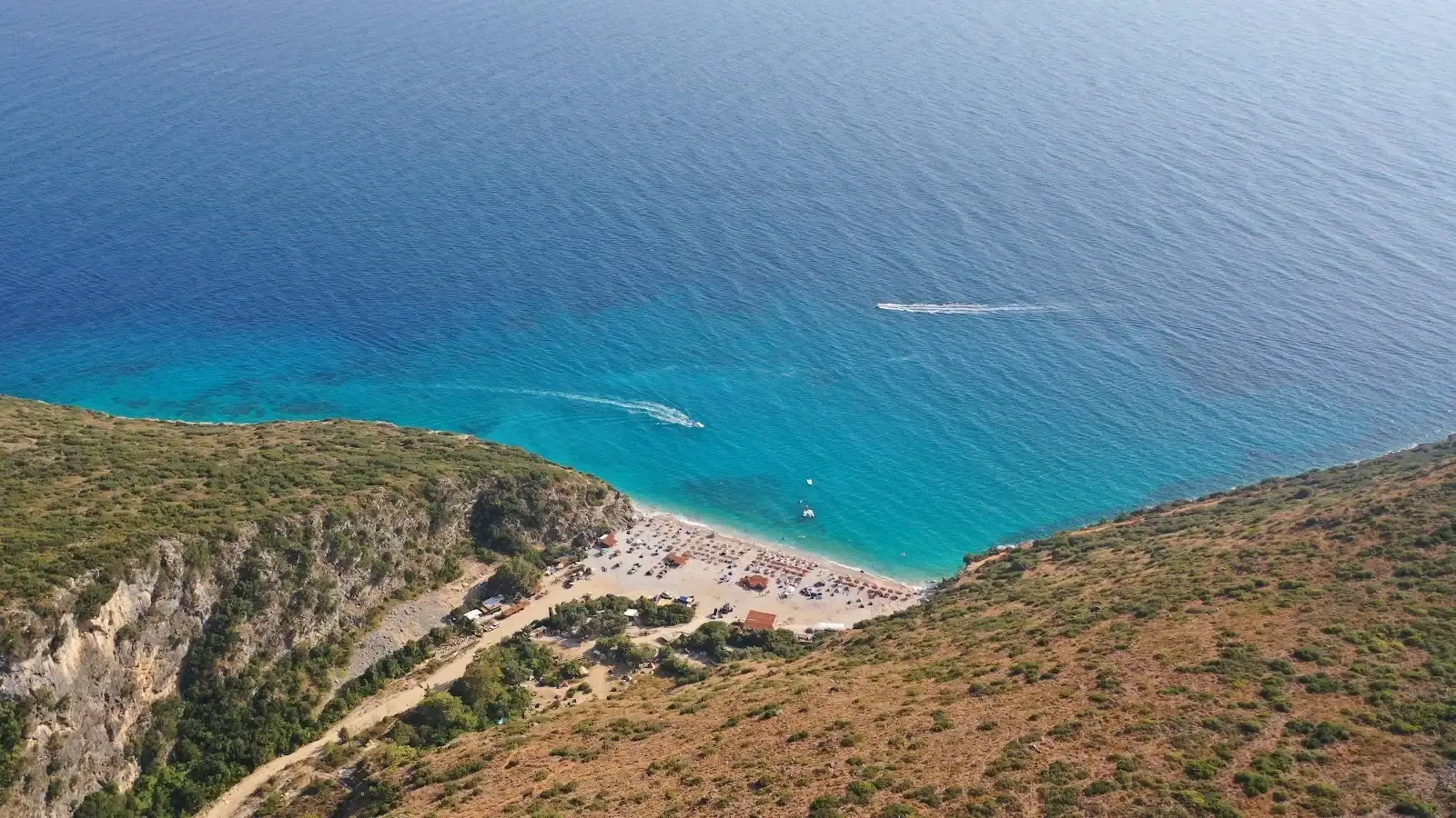 Aerial view of a speedboat crossing a turquoise cove on the Albanian Riviera, its wake trailing behind it toward a pale beach below scrub-covered headlands