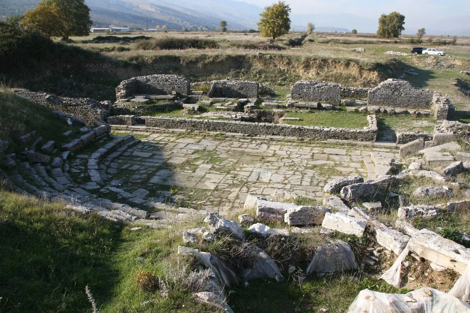 The semicircular ancient Roman stone theatre of Hadrianopolis in a green Drino-valley field with mountains behind