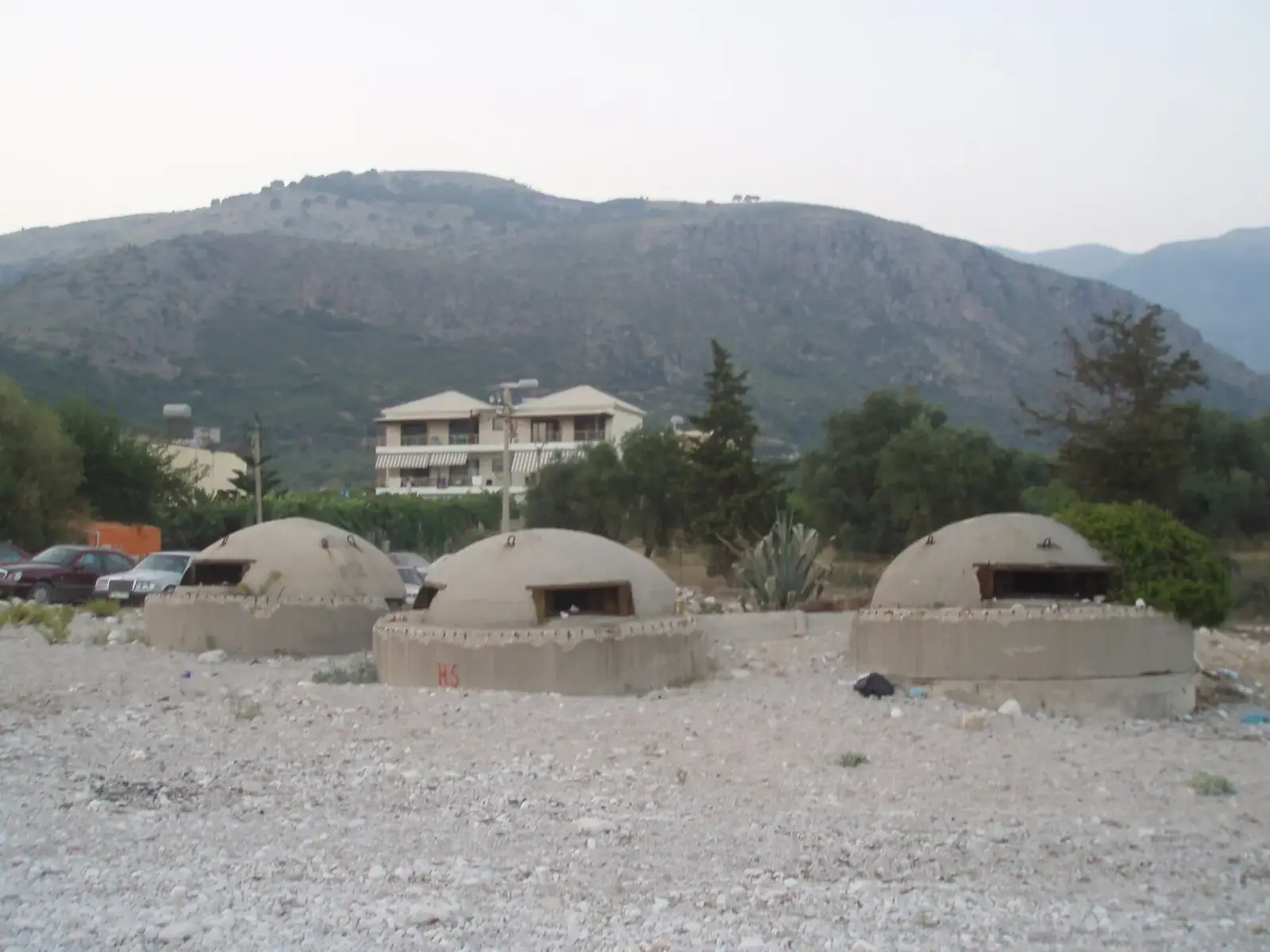 Three concrete dome bunkers standing in a row on the shingle at Himara, with houses and the Ceraunian hills behind them.