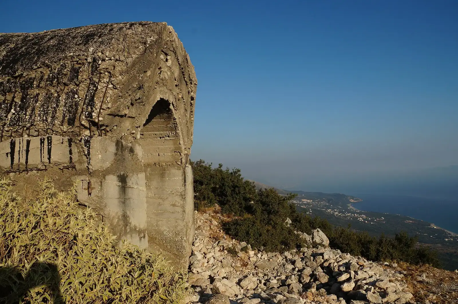 Large weathered concrete bunker with an embrasure and exposed reinforcing bars on the ridge above the Llogara pass, the Riviera coastline far below.