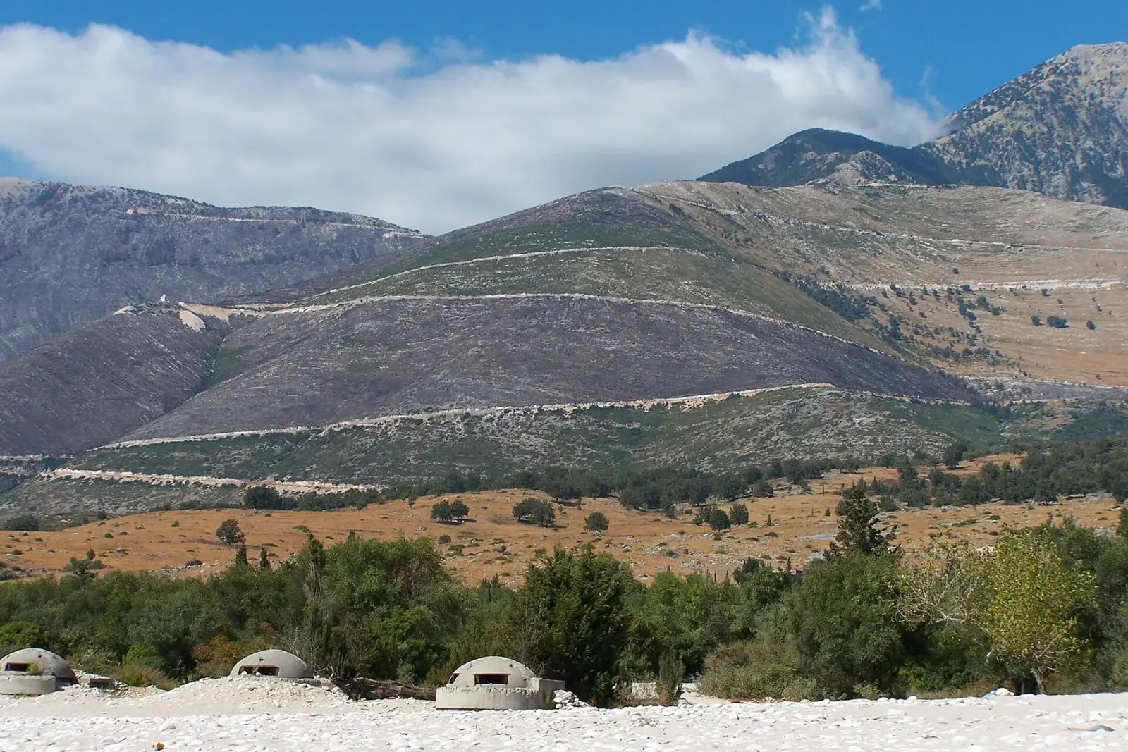 Three dome bunkers spaced along a white pebble shore on the Albanian Riviera, with the bare Ceraunian mountains rising behind.