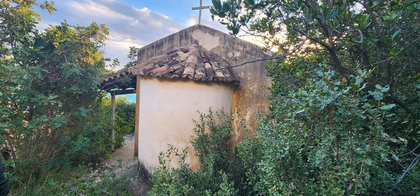 Small whitewashed Orthodox chapel of Saint Nicholas with a stone-tiled roof and cross, surrounded by greenery near Dhërmi