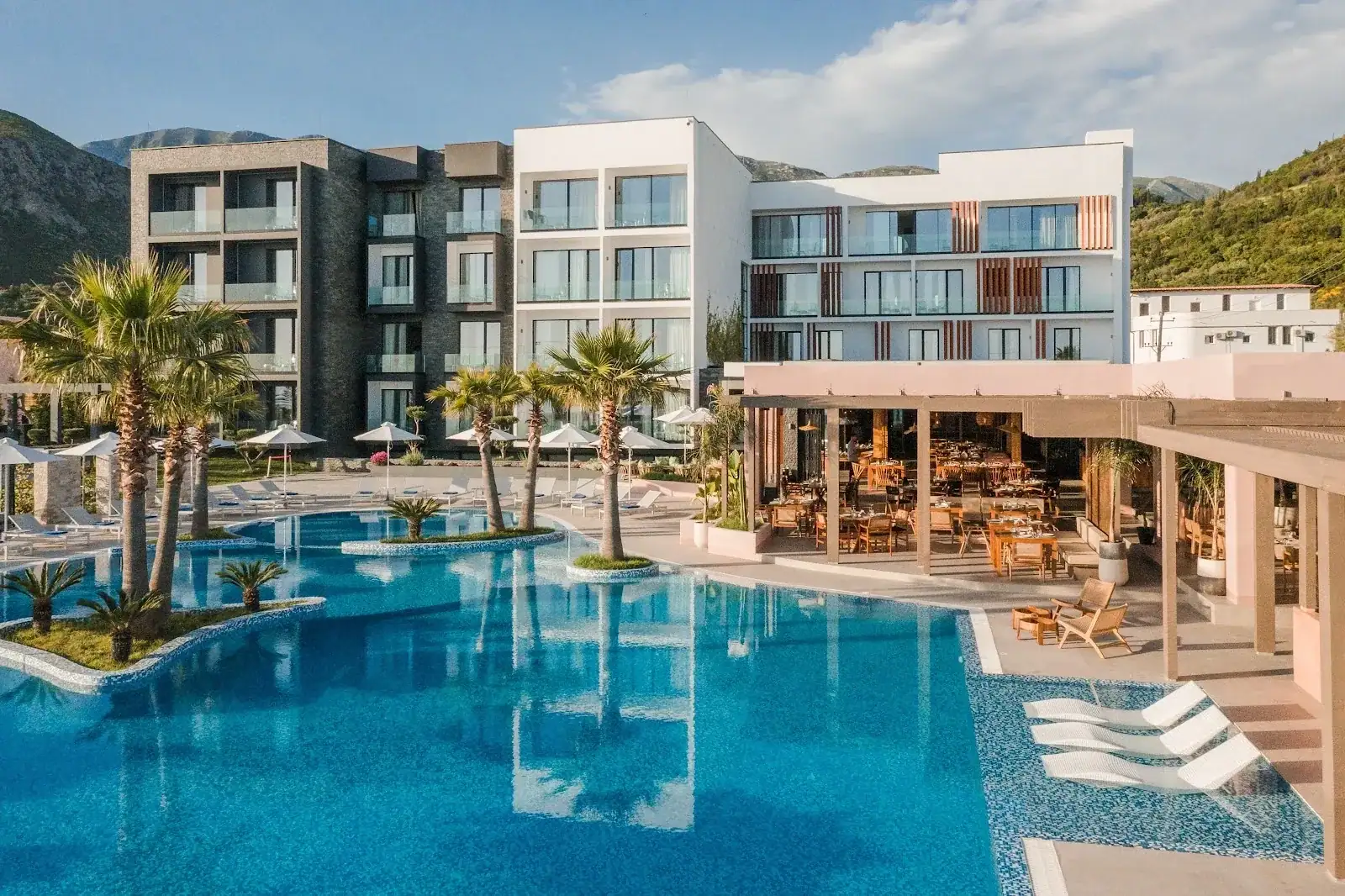 The white and blush façade of Miamar Luxury Hotel behind its outdoor pool, palms and restaurant terrace at Livadh, Himarë