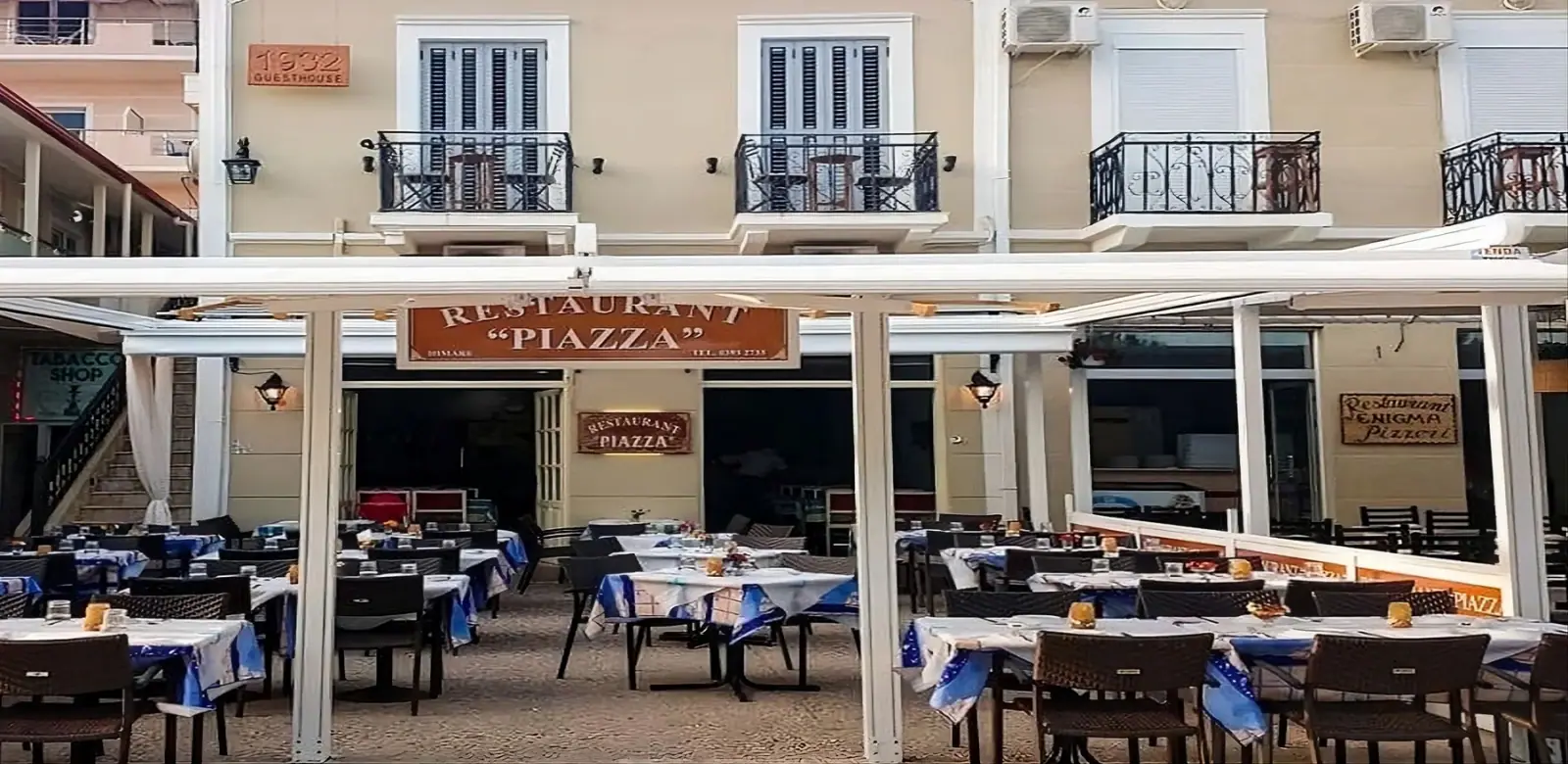 The frontage of Restaurant Piazza in Himarë, the terracotta sign hanging under the white awning above laid tables