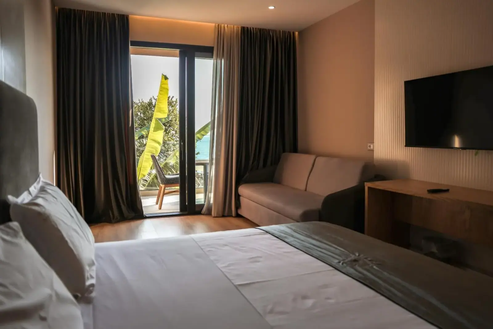 A room with a white bed, a sofa, a wall-mounted television and a glazed door onto a balcony of banana leaves