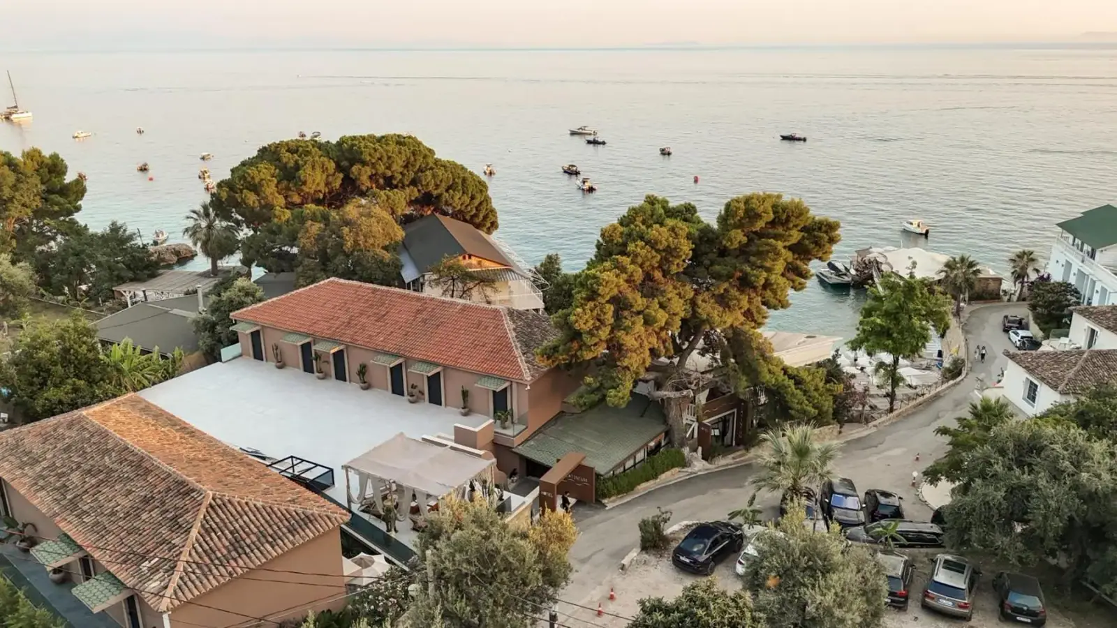 A drone view over tiled roofs and umbrella pines to a turquoise bay with small boats moored offshore
