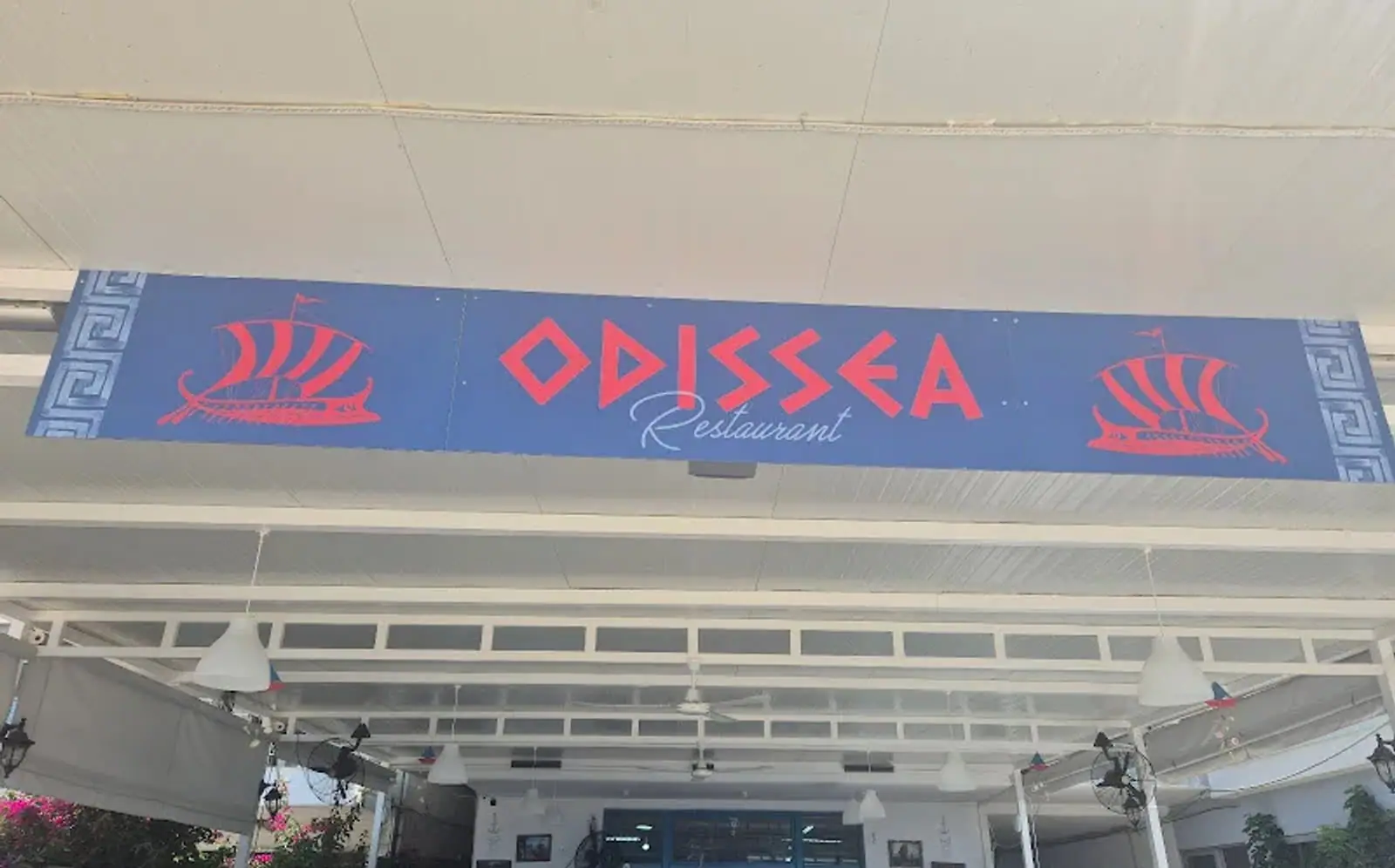 The painted Odissea Restaurant sign, red lettering and two galleys on a blue board with a Greek key border, fixed under the white pergola roof