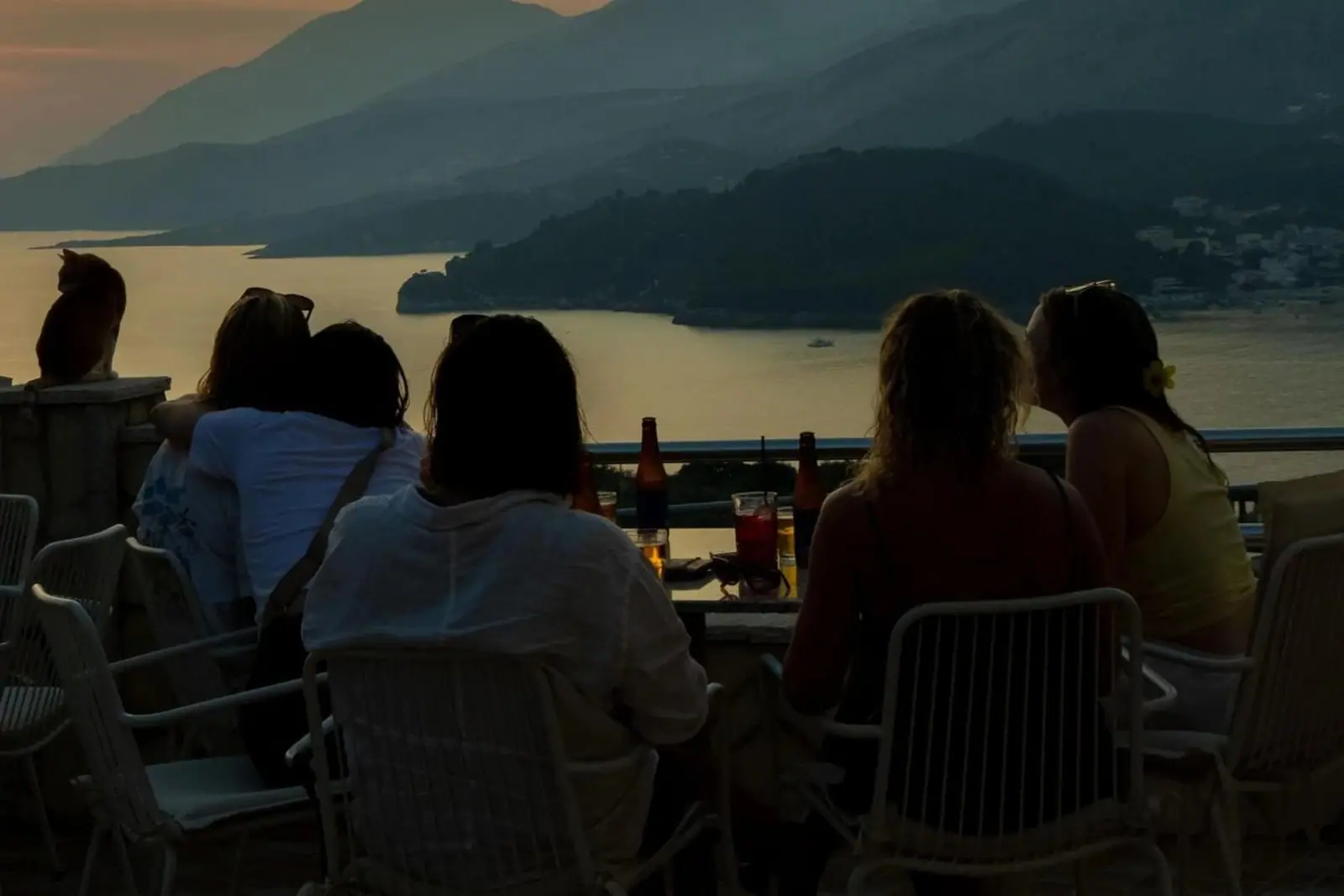 Guests at the terrace rail above Himarë bay watching the sun drop towards the sea, drinks on the table