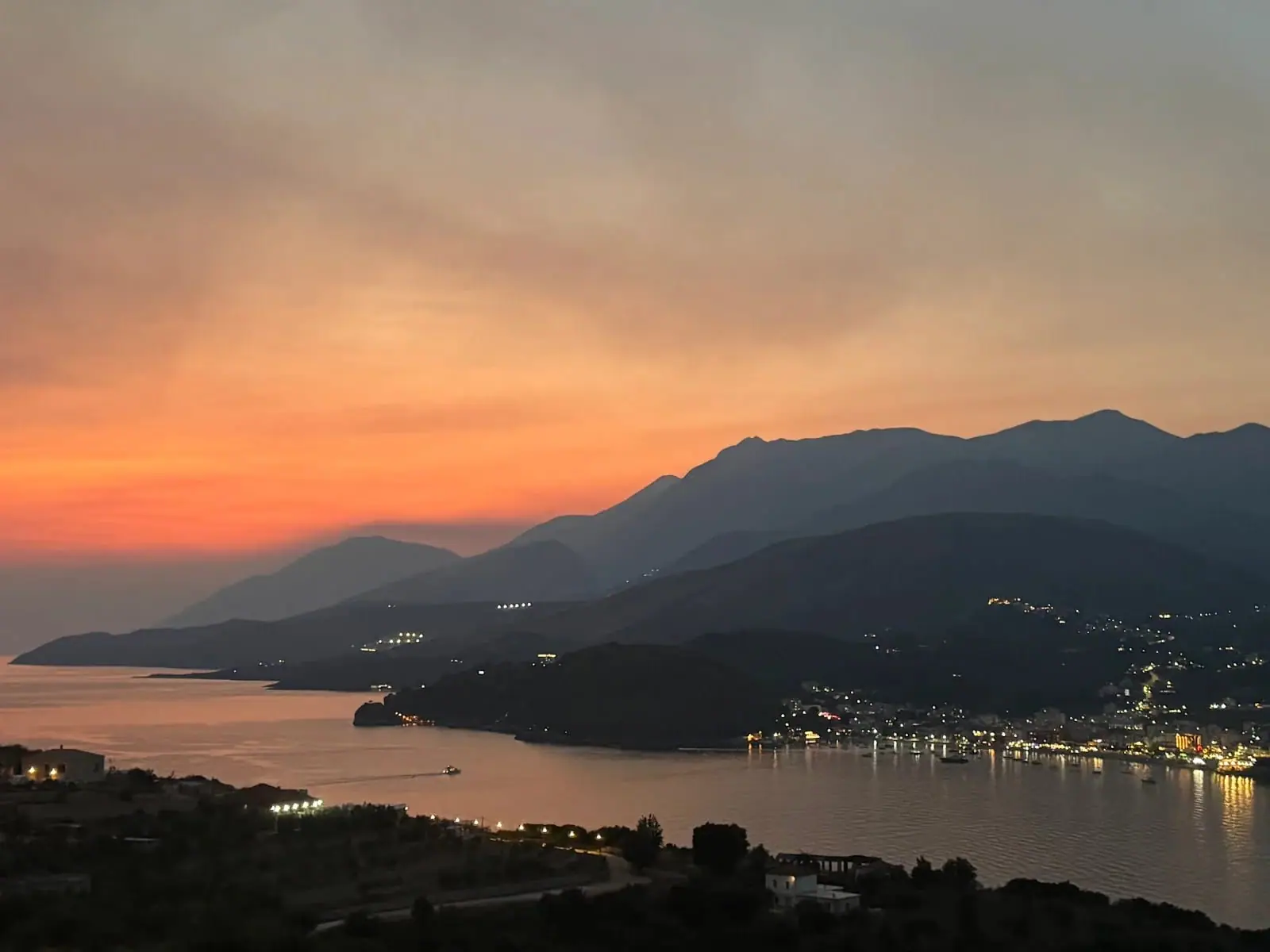 Himarë bay from the terrace just after sunset, orange banding the sky over dark headlands and the town lit along the shore