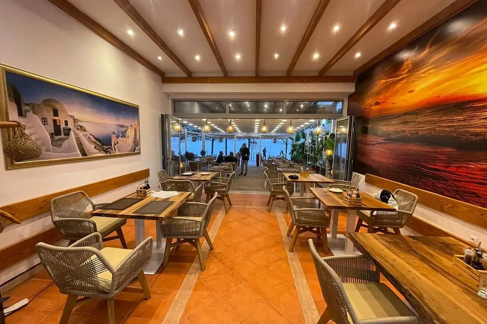 The indoor dining room, terracotta tiled floor and timber-beamed ceiling, opening through folding glass onto a lit terrace and the sea