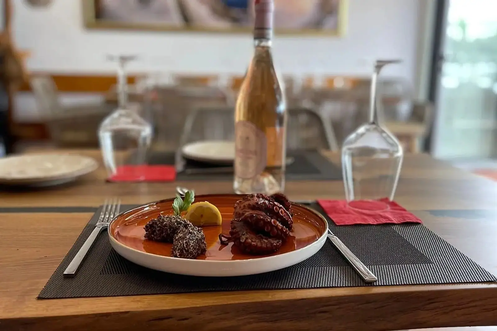 Grilled octopus with lemon and two dark croquettes on a copper-glazed plate, a bottle of rosé and upturned glasses behind it on a timber table