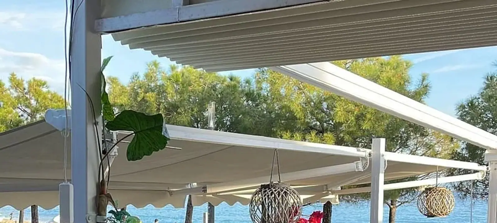 Retractable awnings and a pergola over the terrace, woven rattan lanterns hanging beneath them, pines and the Ionian behind