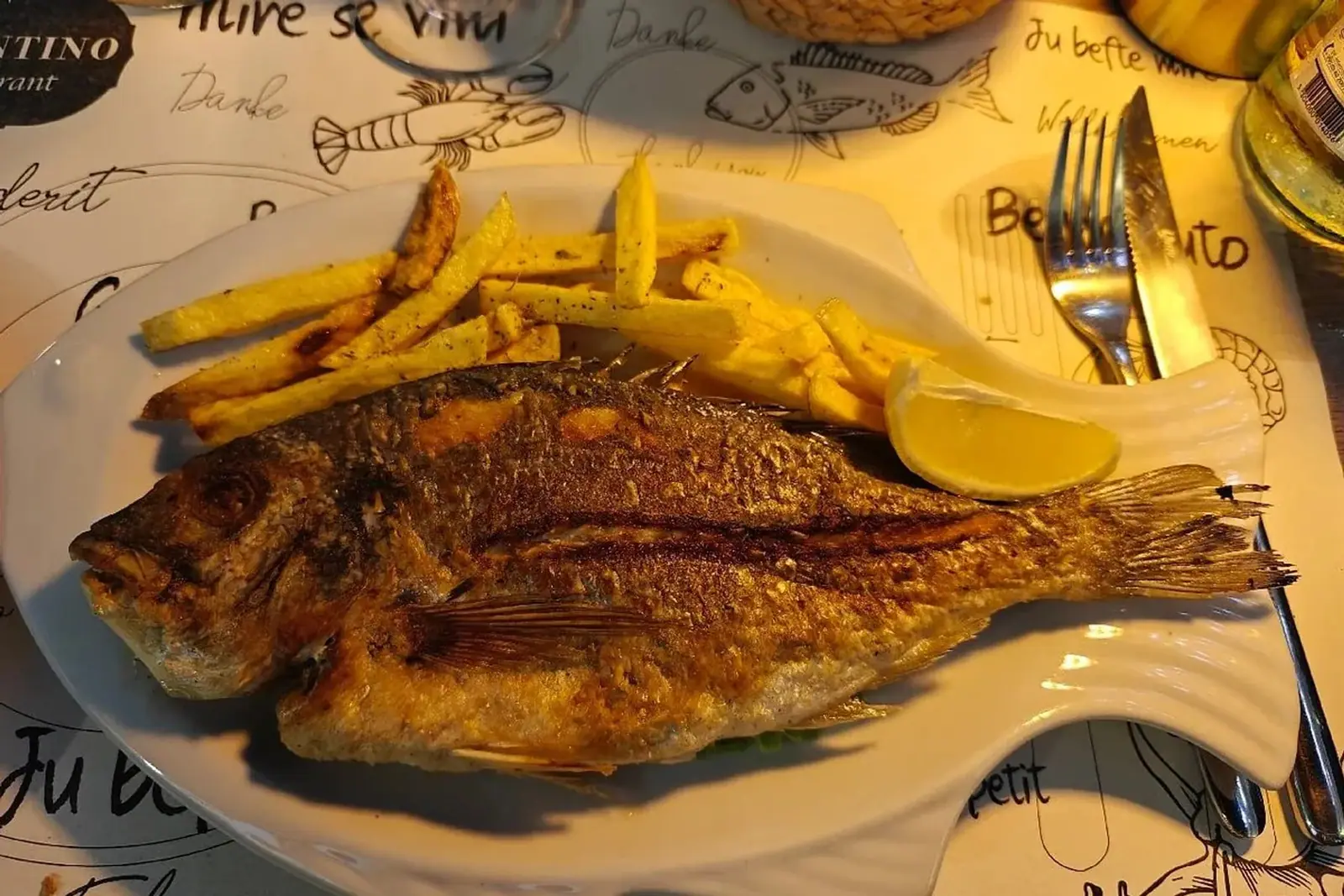 A whole fish, crisp-skinned, on a fish-shaped white plate with chips and a wedge of lemon, on a placemat printed with the Valentino Restaurant mark