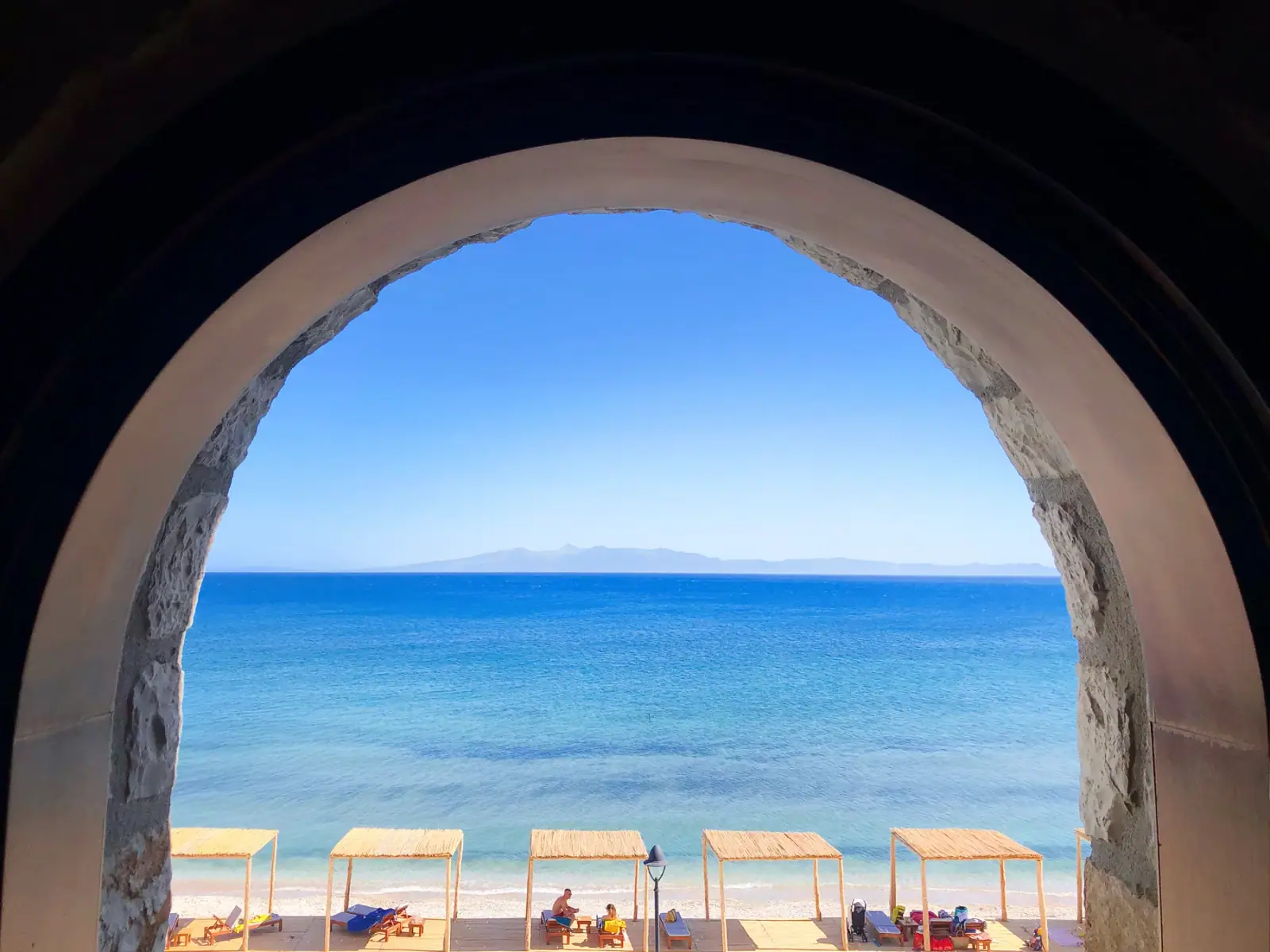 A round stone arch opening straight onto the bay, with flat thatched shade canopies in a line on the pebbles below