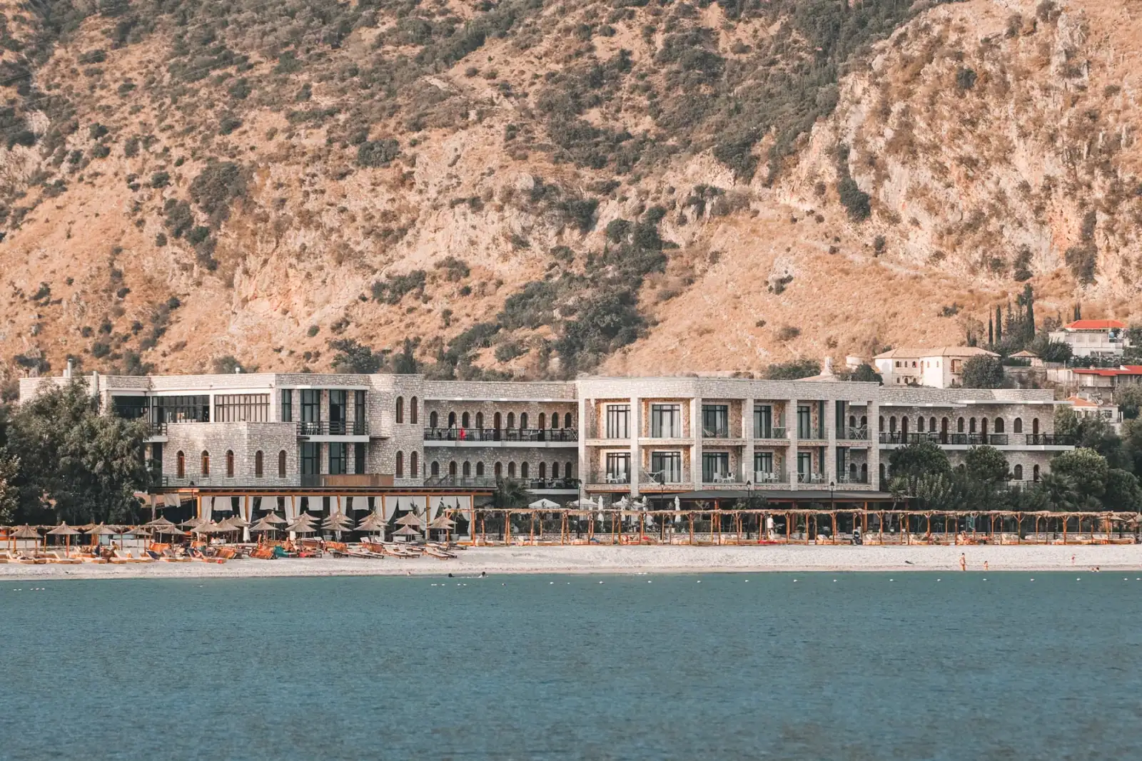 The long limestone front of Valta Hotel photographed from the water, its arched windows in rows, round thatched parasols on the pebbles below and a scrub-covered mountain behind