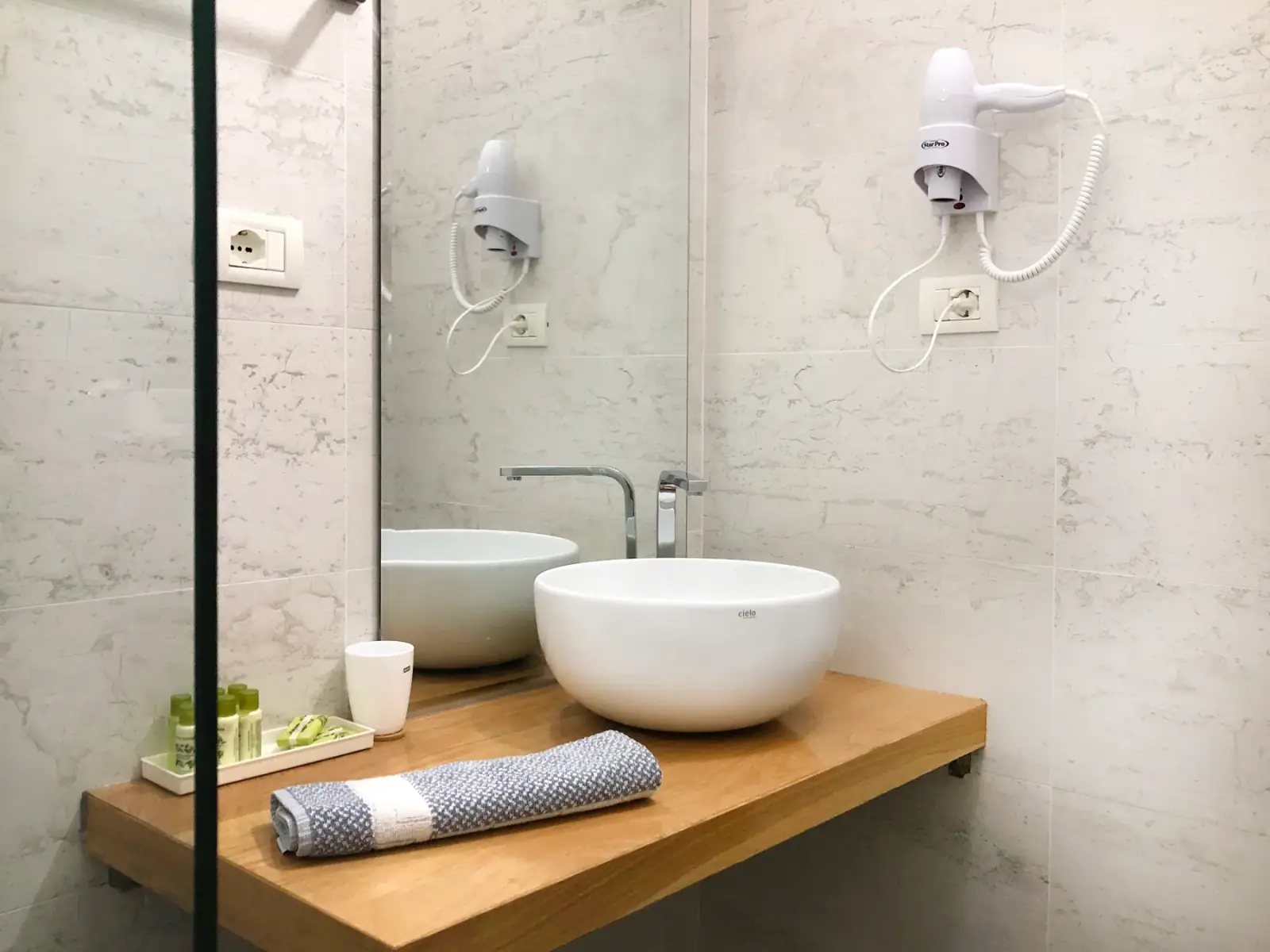 A round bowl basin on a timber shelf below a full-height mirror in a marble bathroom, a hairdryer on the wall