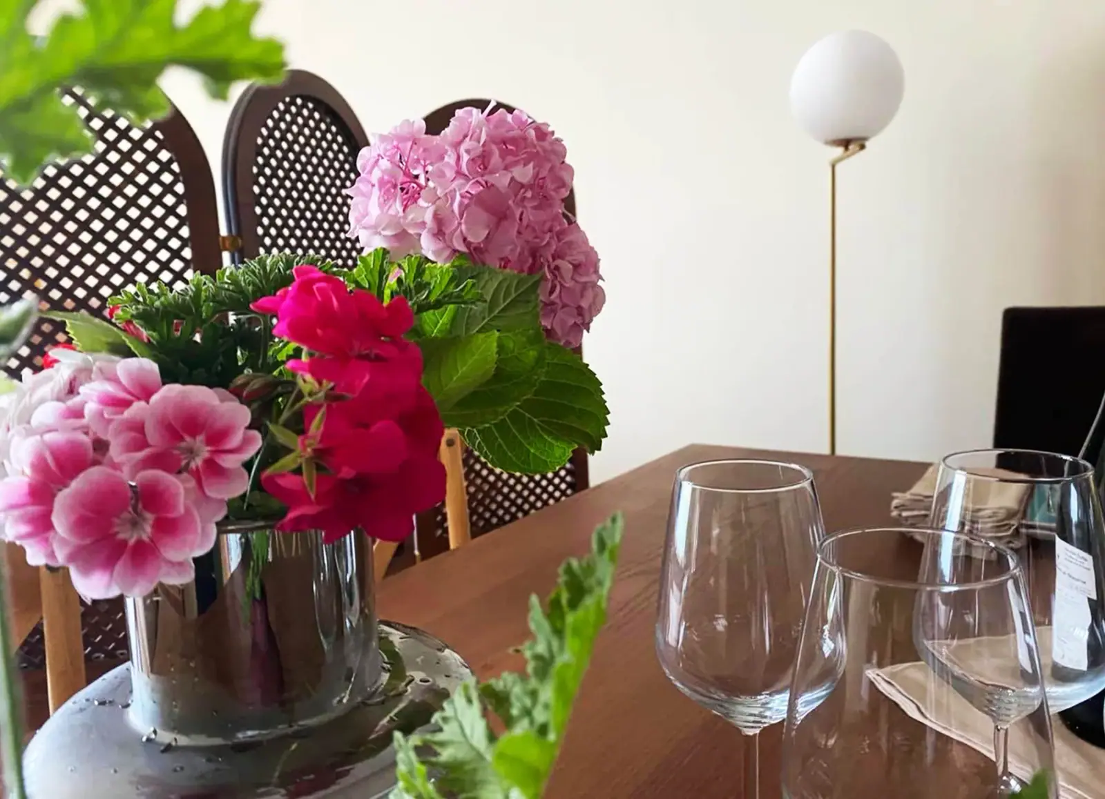 Three wine glasses and a glass vase of pink hydrangeas and geraniums on a dark timber table, lattice-backed chairs around it