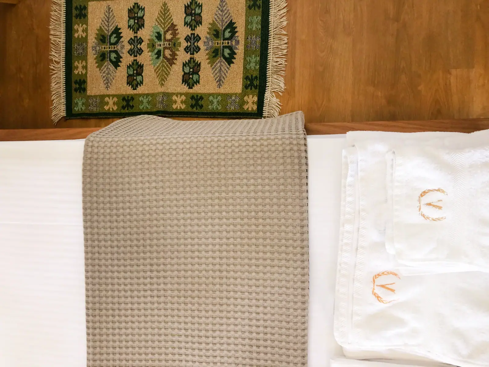 Folded white towels on a made bed beneath a green woven kilim hung on the timber headboard wall
