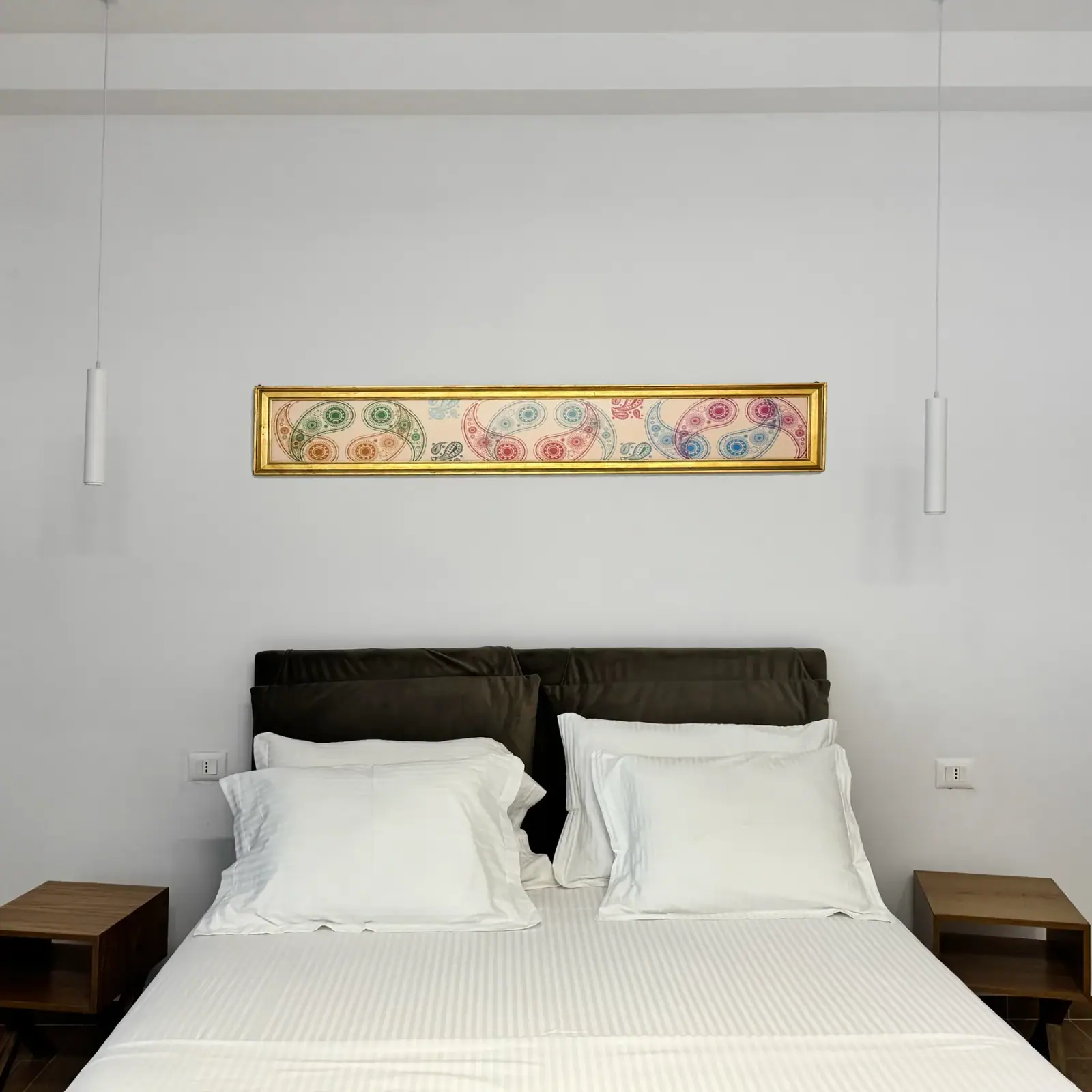 A double bed with a dark upholstered headboard beneath a long framed panel on a white wall