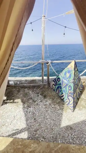 A large hand-painted panel in blue, green and cream propped against the rope railing at Albania Glamping, seen from inside a tent doorway with festoon lights overhead and pebble ground below