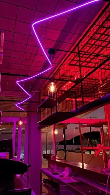 A violet neon zigzag running across the terrace ceiling at night, with a caged filament lamp and the dark sea beyond