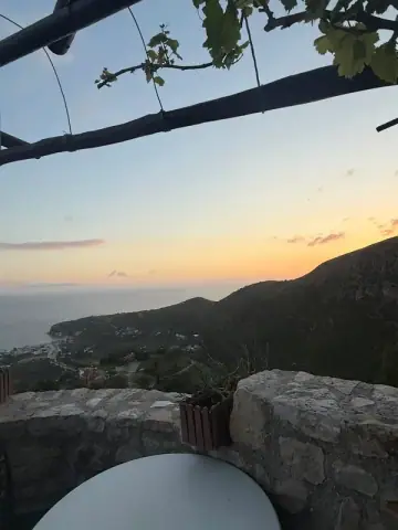 Dusk from the parapet at Café Butterfly — the sky apricot above a flat grey sea, a wooded ridge running out to the right, houses scattered on the slope below, two peeled roof poles and a vine crossing the top of the frame