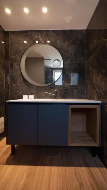 A bathroom lined in bronze-brown marble-effect tile with a round black-framed mirror, a navy vanity unit with an open oak cubby, and a wood-look plank floor