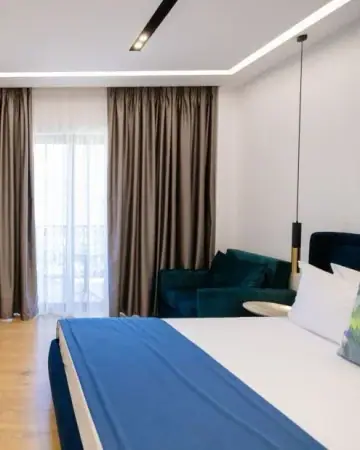 A double room at Chimaera Hotel with a blue bed runner, a teal armchair and floor-length curtains at the balcony door