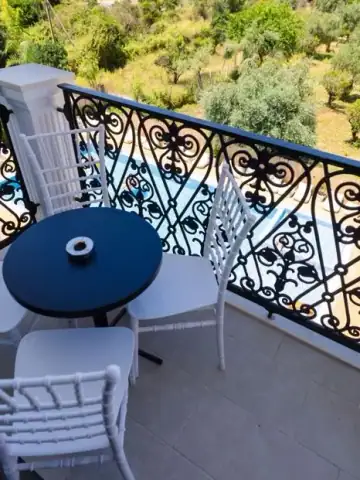 A balcony at Chimaera Hotel with a dark round table and white chairs behind a scrolled wrought-iron rail, the pool and an olive terrace below