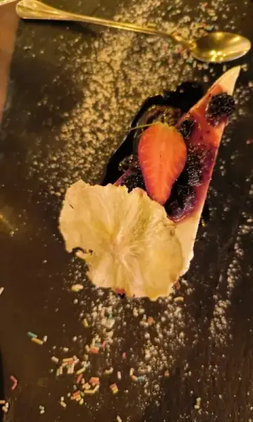 A wedge of pale set dessert under dark berry compote and a whole blackberry, with a halved strawberry and a large dried pineapple slice, on dark slate strewn with sugar and coloured sprinkles beside a gold spoon