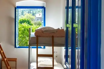 A bunk bed beside an open blue-framed window at Himara Hostel, with garden greenery outside