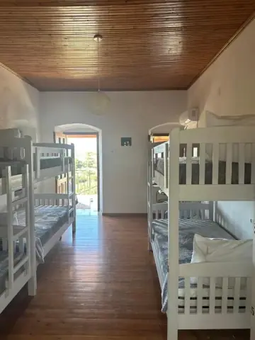 A dormitory with white bunk beds on both sides and an open doorway at the end leading out to a balcony