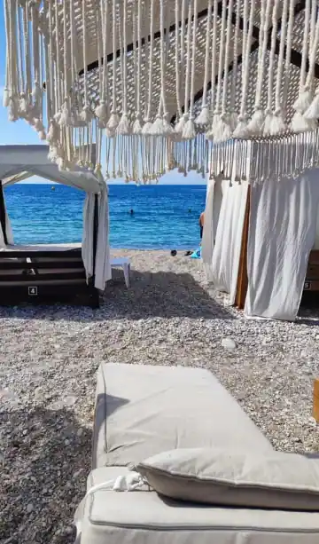 A white-curtained four-poster daybed at the top of the pebbles, with the open sea beyond