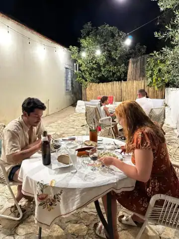 Guests eating at a table in a walled courtyard at night under a string of lights, with a bamboo screen and an olive tree behind them
