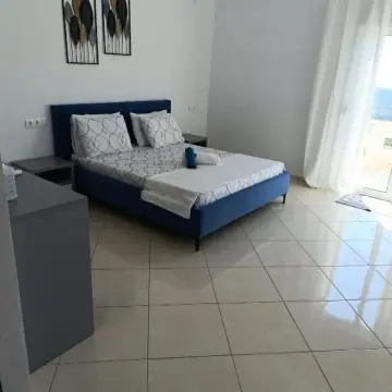 A blue upholstered double bed under two framed leaf prints in an apartment at Nikolari Resorts, cream floor tiles running to a curtained balcony door