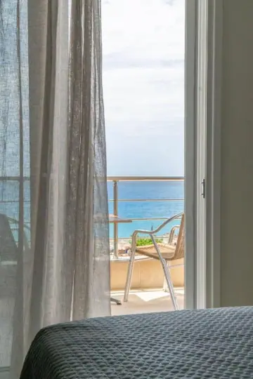 An open balcony door seen from the foot of a bed, a linen curtain drawn back, a wicker chair and the sea beyond the rail