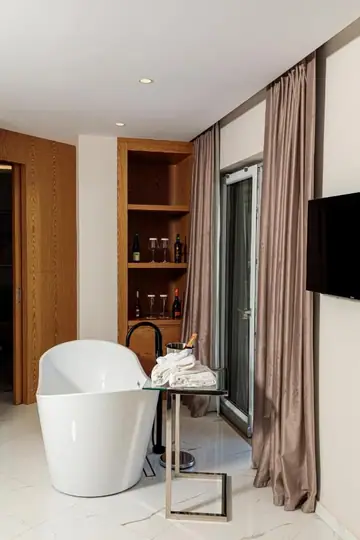 A freestanding white bath with a black floor-standing tap in a suite, beside a full-height glazed door with floor-length drapes, a glass side table with folded towels, and a recessed oak niche holding wine glasses and bottles