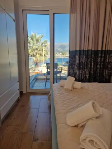 A sliding balcony door in one of the rooms, a black mesh chair on the balcony, a palm, and the bay towards Himarë beyond