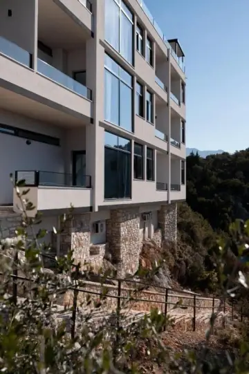 The white rendered flank of Wait 'n Sea on its rocky slope, stone-clad at the base, with black-framed glass and cantilevered balconies above and the mountains behind