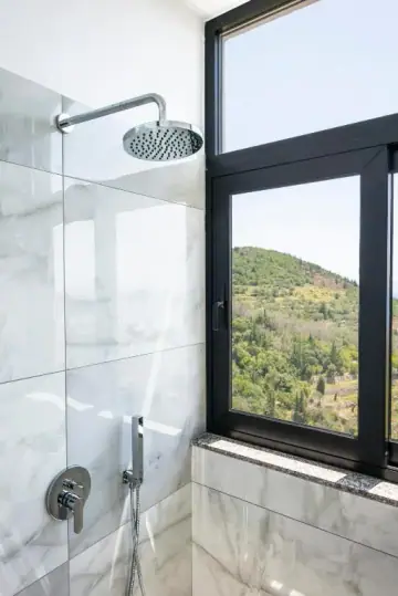 A rain shower behind a glass screen in a white marble bathroom, beside a black-framed window looking out onto the wooded hillside