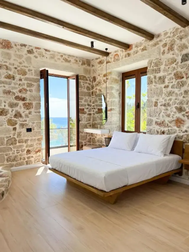 A wide bed on a low timber platform, rubble limestone on every wall in the frame, a glazed door standing open onto a balcony above the sea, and a two-leaf window onto green.