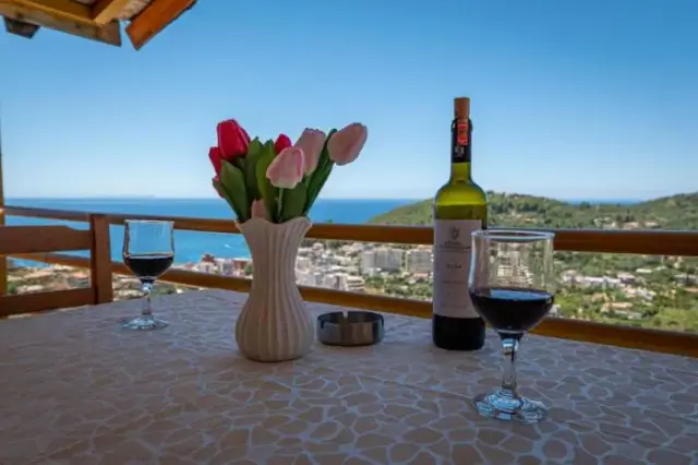 A laid table under a timber canopy on the terrace, with Himarë town and the Ionian spread out below the rail
