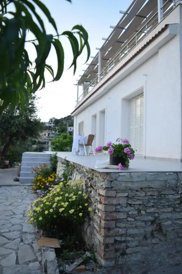 A white house with shuttered windows above a stone terrace with a wicker chair and pots of geraniums, the village below
