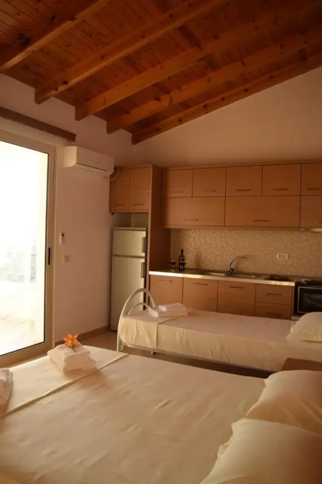 A fitted kitchen of pale wood units with a worktop, sink and oven, a bed in the same room and an air conditioner above