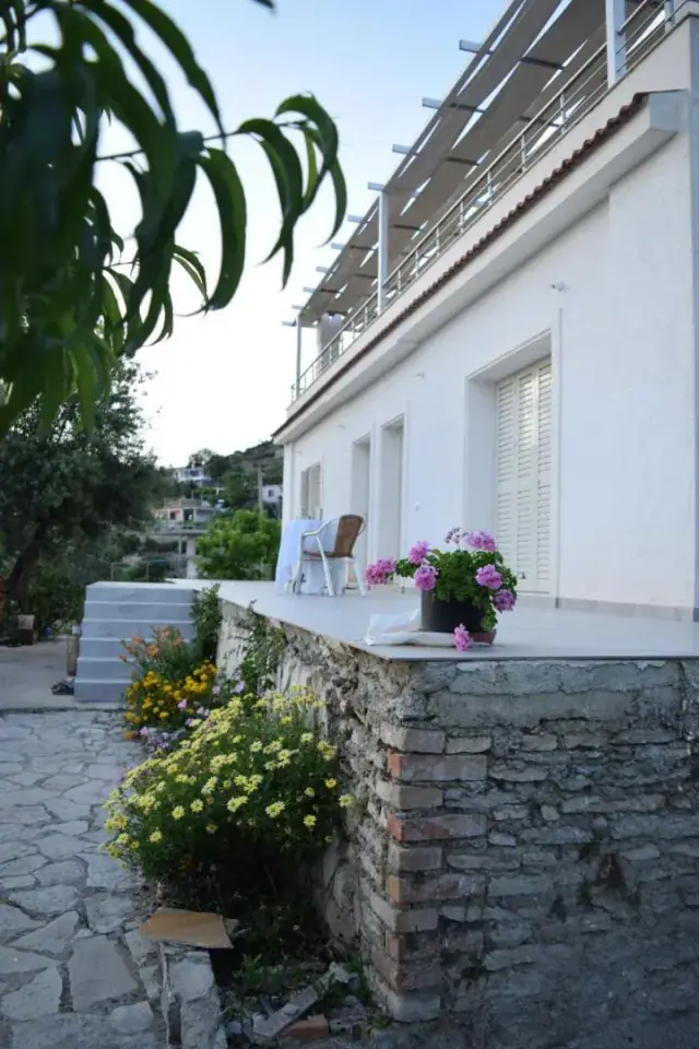 A white house with shuttered windows above a stone terrace with a wicker chair and pots of geraniums, the village below
