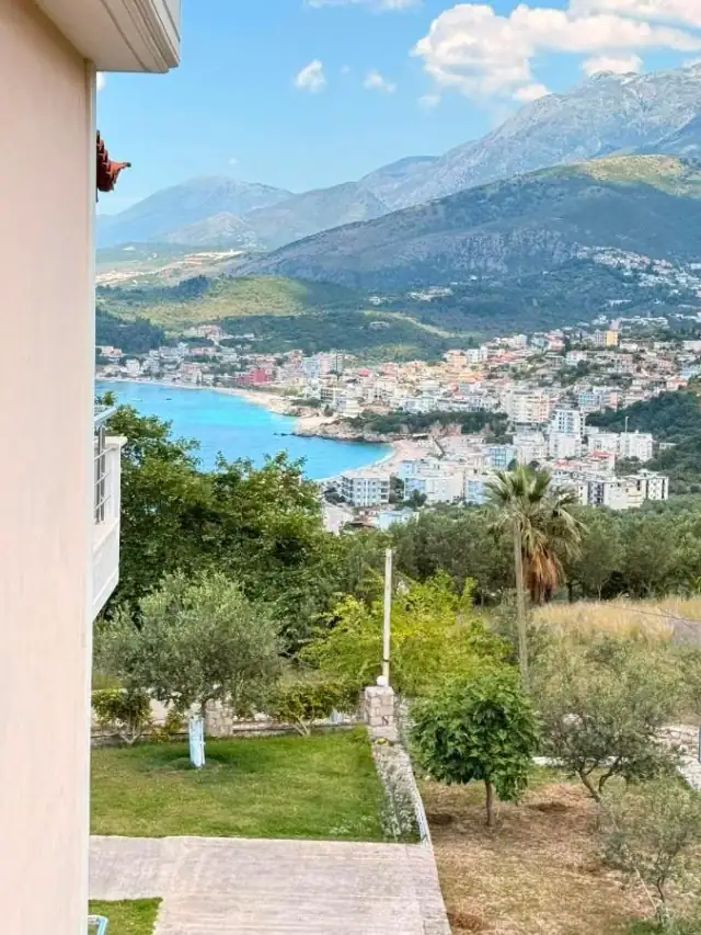 Himarë town and its turquoise bay seen over the property's garden from the floor above it, with the mountain wall rising behind the town