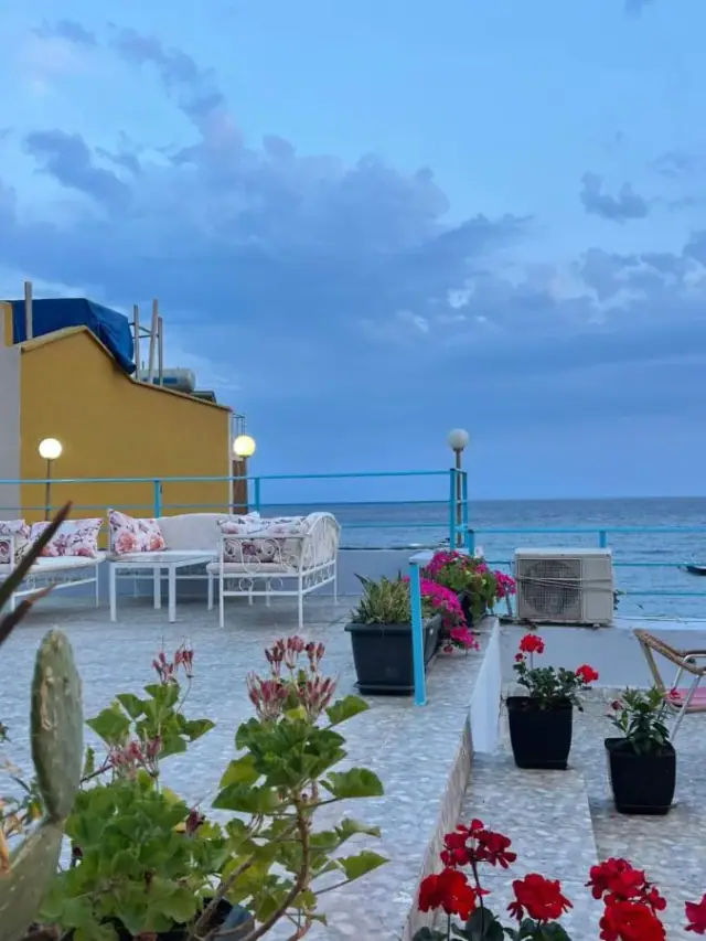 The hotel roof terrace at dusk, with turquoise railings, white seating, potted geraniums and the open sea beyond.